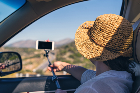 Blogger girl in the straw hat taking selfie picture or video using smartphone and selfie stick inside of car. Holidays and tourism conceptの写真素材
