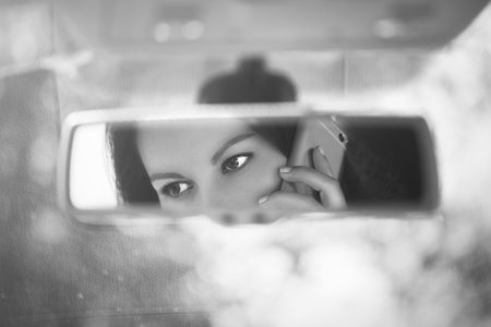 Black and white art picture of young woman talking on a mobile phone in the car rear view mirror.の写真素材
