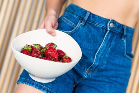 Bottom view on women in denim shorts with ripe strawberry in the bowl on the background of wooden beamsの写真素材