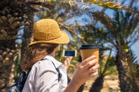 Back view on the Woman in the straw hat with paper cup of coffee taking selfie picture or video using smartphone in the palm forest.の写真素材