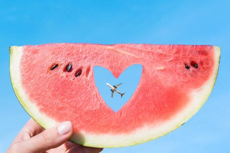 Ripe piece of watermelon with heart shape hole in female hands on the background of the blue sky and plane. Summer Conceptの写真素材