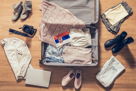 Woman's clothes, laptop, camera, russian passport and flag of Serbia lying on the parquet floor near and in the open suitcase. Travel conceptの写真素材
