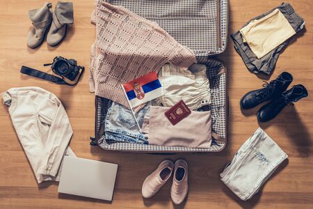 Woman's clothes, laptop, camera, russian passport and flag of Serbia lying on the parquet floor near and in the open suitcase. Travel conceptの写真素材