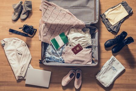 Woman's clothes, laptop, camera, russian passport and flag of Nigeria lying on the parquet floor near and in the open suitcase. Travel conceptの写真素材