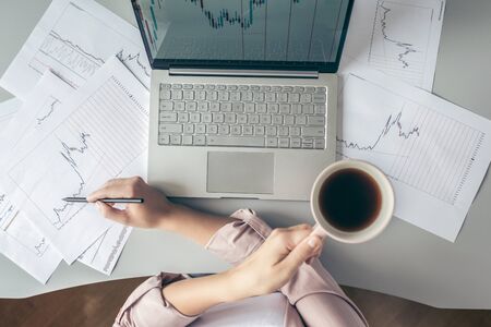 Top view. Young business woman sitting at table with cup of coffee and working on the laptop with graphics and charts printed on the paper.の写真素材