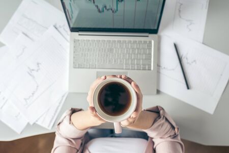 Top view. Young business woman sitting at table with cup of coffee and working on the laptop with graphics and charts printed on the paper.の写真素材