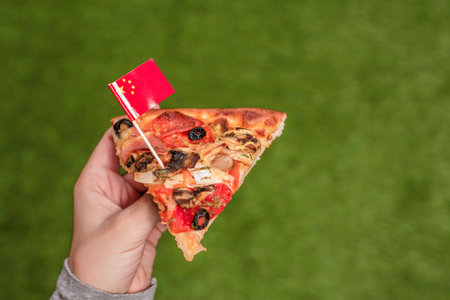 A slice of pizza in female hands with Chinese flag in the form of a toothpick. Lunch on the green...