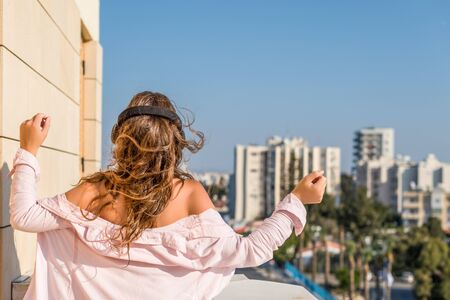 woman in a pink shirt on the balcony dancing and listening music in a headphones against the city backgroundの写真素材
