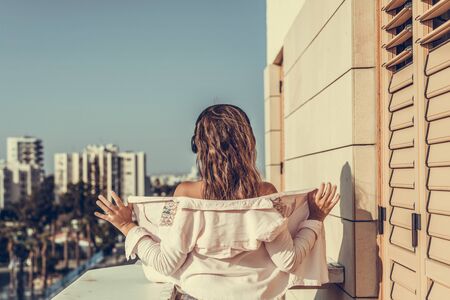woman in a pink shirt standing on the balcony and listening music in a headphones against the city backgroundの写真素材