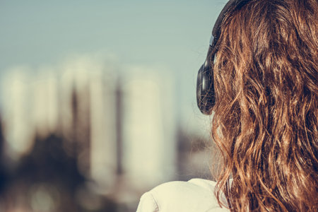 woman in a pink shirt standing on the balcony and listening music in a headphones against the city...