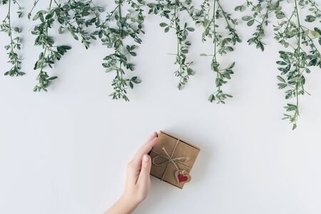 Woman packing gift in a box of kraft paper on white wood table. Green leaves, branches. concept. Flat lay, top view. の写真素材