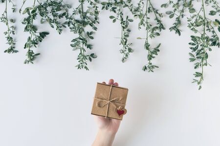 Woman packing gift in a box of kraft paper on white wood table. Green leaves, branches. concept. Flat lay, top view. の写真素材