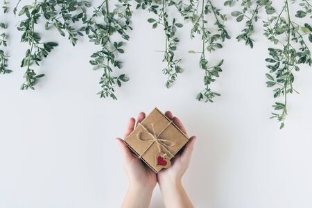 Woman packing gift in a box of kraft paper on white wood table. Green leaves, branches. concept. Flat lay, top view. の写真素材