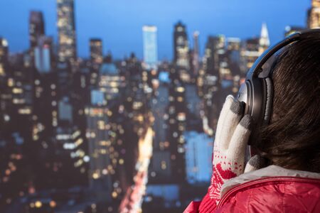 young woman in a winter jacket and knitted gloves listening music in the headphones on the roof against the backdrop of night New York. Aerial and panorama view of skyscrapers of New York City. Top view of midtown of Manhattan. の写真素材