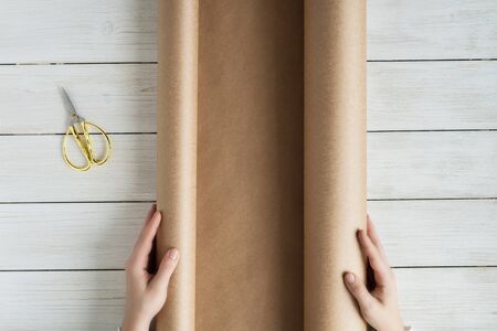Female hands unfolding a roll of wrapping brown craft paper for packing gifts and flowers on the wooden white table. Copy space.の写真素材