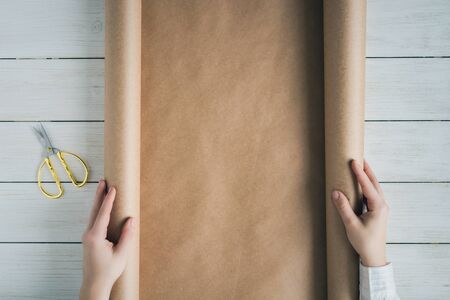 Female hands unfolding a roll of wrapping brown craft paper for packing gifts and flowers on the wooden white table. Copy space.の写真素材