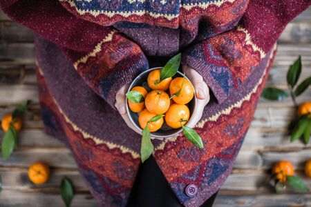 Woman in a huge winter sweater sits on the wooden rustic floor with small iron bucket of tangerines (mandarin).の写真素材