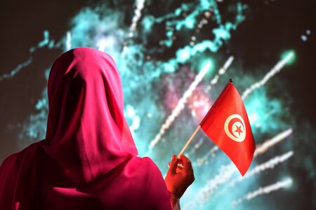 Muslim woman in a scarf holding flag of Tunisia during fireworks at night.の写真素材