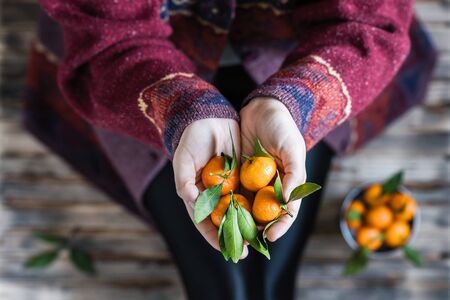 Woman in a huge winter sweater sits on the wooden rustic floor with small iron bucket of tangerines (mandarin).の写真素材