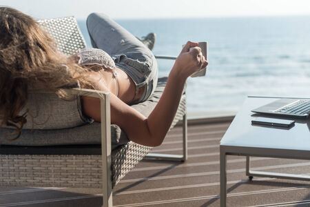 Beautiful brunette woman freelancer in a swimsuit and jeans resting on a couch and sunbathing on the terrace with a cup of coffee at summer day. Sea view. WFH. Working from home, laptopの写真素材