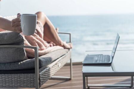 Beautiful brunette woman freelancer wrapped in a plaid resting on a couch and sunbathing on the terrace with a cup of coffee at summer day. Sea view. WFH. Working from homeの写真素材