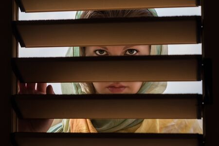 Lonely orthodox young woman in a headscarf looks through the wooden blinds. Conceptの写真素材