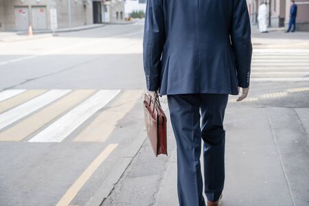 Alone businessman in a blue suit with briefcase in a protective white rubber gloves on the crosswalk on the city street. Health protection against viruses, germs and bacteria. Coronavirusの写真素材