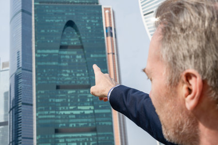Alone middle-aged gray-haired businessman in a blue suit showing by finger on the skyscrapers of Moscow City.Low angle view. Russiaの写真素材