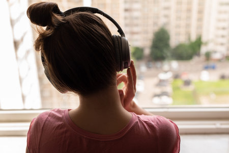Back view of a lonely young woman listening to music with headphones standing at the window of her...