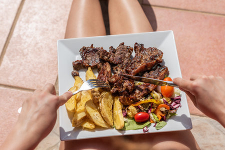 One woman having a lunch putting plate with lamb potato and salad on the knees and eating outside on...