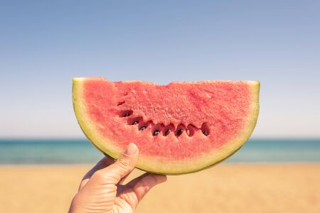 Bitten ripe piece of watermelon in woman hands on the beach in a hot summer day. Conceptの写真素材