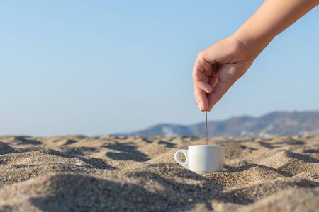 Woman drinking coffee from a little cup and holding a small spoon on sunny sand beach opposite sea wave. summer. take away. breakfastの写真素材