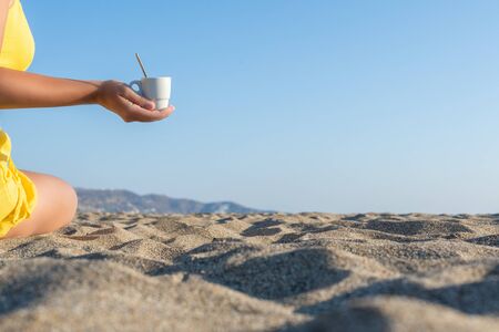 Woman drinking coffee from a little cup and holding a small spoon on sunny sand beach opposite sea wave. summer. take away. breakfastの写真素材