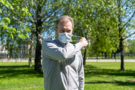 Happy middle-aged gray-haired man taking off a medical mask from his face in the spring park. Over...