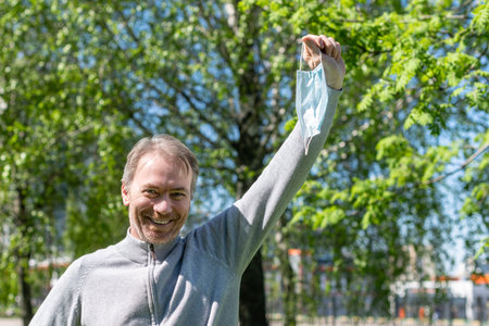 Happy middle-aged gray-haired man taking off a medical mask from his face in the spring park. Over...