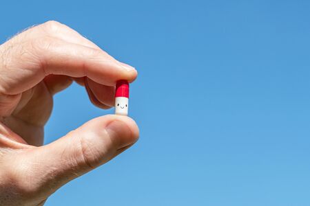 A man holding in his hand a funny smiling pill on the blue sky background. The cure for viruses, bacteria, allergies. Creativeの写真素材