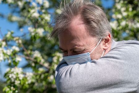 A white-haired middle-aged man in a medical mask sneezing in a park among flowering trees. Seasonal Allergy. Spring time.の写真素材