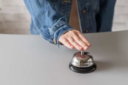 Hand of guest in a denim jacket ringing in silver bell. reception desk with copy space. Hotel...