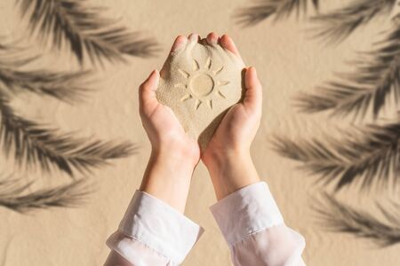 Female hand with a handful of sand with sun hand-drawn among shadow of a palm tree branch of a sandy tropical beach. Holiday concept. Creative, background, travel, summer. Flat layの写真素材