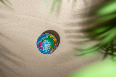 Rubber toy globe on sandy tropical beach under branches of palm and shadow palm tree. Holiday...