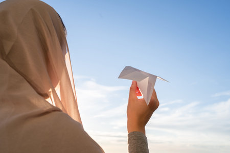 Silhouette of muslim woman in head scarf launching a paper airplane on blue sunset sky background....