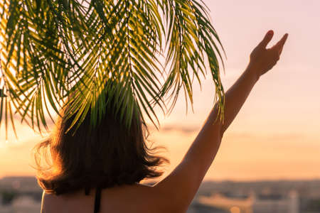 Happy girl stands under a palm tree and waves her hand while welcoming the sunset. Summer, freedom conceptの写真素材