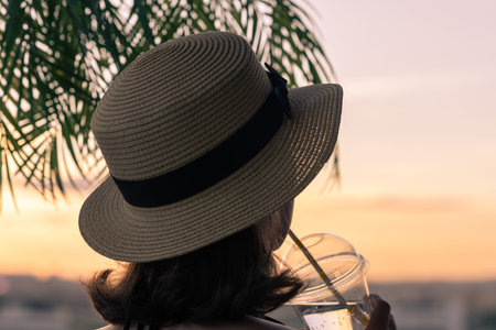 Back view of a beautiful girl with lemon water in a glass in a straw hat against the background of...