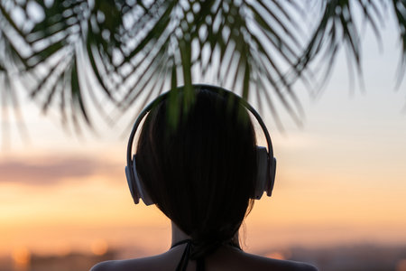 Back view silhouette of relaxed woman wearing headphones meditating listening to music on the beach at sunset in the branches of palm trees.の写真素材