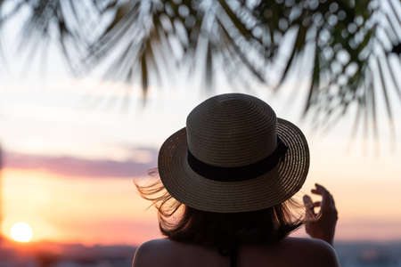 Back view of a beautiful girl in a straw hat against the background of the sea in branches of palm trees. Sunset beach. Summer and freedom concept.の写真素材