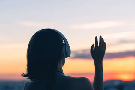 Lonely sad woman in headphones standing opposite the setting sun over the horizon of the sea. Girl waving hand to sunset sky. Goodbye gesture.の写真素材