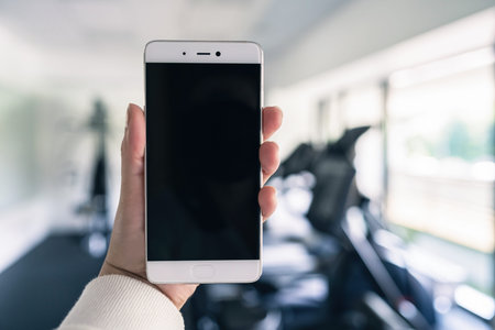 Woman hand with mobile phone in the gym opposite the sport equipment and jogging simulators. Fitness...