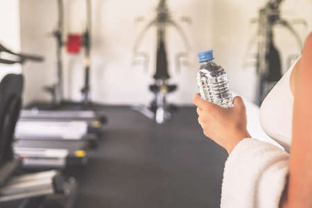 Cropped. Sporty girl with towel on shoulders and bottle still water in the fitness gym opposite the sport equipment and jogging simulators. Resting time. Health concept.の写真素材