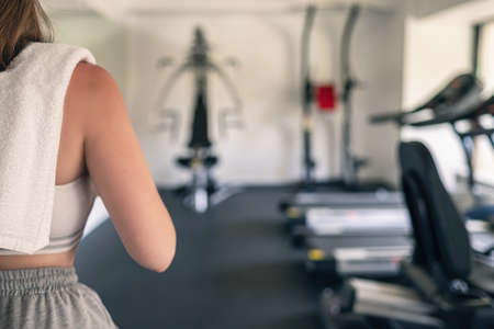 Cropped. Sporty girl with towel on shoulders in the fitness gym opposite the sport equipment and jogging simulators. Resting time. Health concept.の写真素材