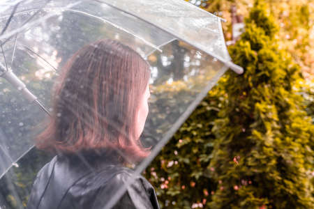 Autumn. Lonely sad redhead woman walking in a park, garden. View through wet transparent umbrella with rain drops. Rainy day landscape. Vintage tonedの写真素材